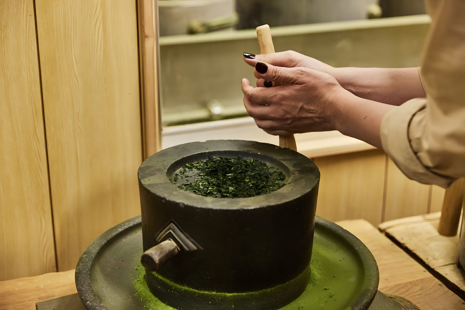 Close-up of a hand grinding green tea leaves with a wooden pestle in a traditional stone mortar, set in a warm wooden tea preparation area
